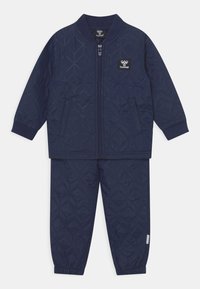 Marinblå kviltad träningsoverall med zipjacka och elastiska byxor. Har diamantmönster, ribbade mudd och sidofickor.