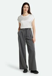 Pantalones de pierna ancha grises con plisados, cintura con cordón y bolsillos grandes, combinados con una blusa ajustada de manga corta blanca. Zapatos negros gruesos.