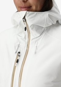 Vrouw in een witte jas met een hoge kraag en beige ritsdetails, die iets glimlacht voor een effen achtergrond.