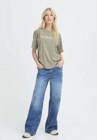 Camiseta verde oliva con "amore" en letras reflectantes, combinada con jeans de mezclilla azul de pierna ancha y zapatillas de color claro.