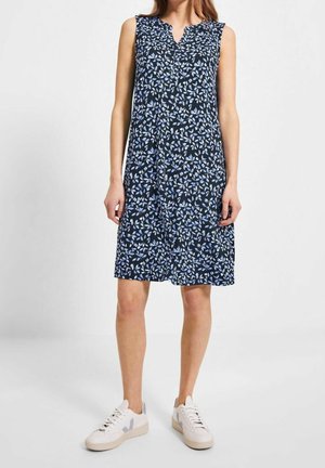 Robe bleu marine sans manches longueur genou avec motif de feuilles bleu clair portée avec des baskets blanches sur une femme debout.