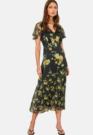 Vrouw draagt een zwarte midi-jurk met geel bloemenprint, doorschijnende fladdermouwen en een V-hals, gecombineerd met zwarte open-toe hakken.