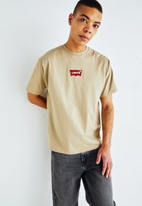 Beige bomulds t-shirt med et rødt broderet Levi's-logo på brystet. Loose fit med korte ærmer og rund halsudskæring.