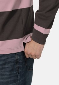 Langärmliges gestreiftes Shirt mit rosa und dunkelbraunen Farben, aus Baumwolle gefertigt. Verfügt über einen seitlichen Schlitz und einen lässigen Schnitt, mit einer Hand in der Tasche.