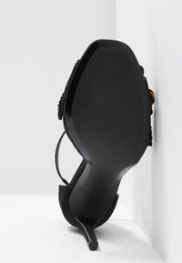 Chaussure noire à talon haut avec bout pointu, talon fin et bride à la cheville, vue de la semelle sur un fond blanc et un mur.