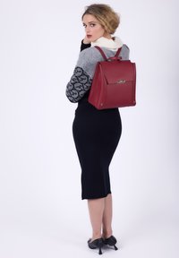 Donna in gonna nera e maglione grigio che indossa uno zaino rosso, volta le spalle con uno sfondo bianco semplice.