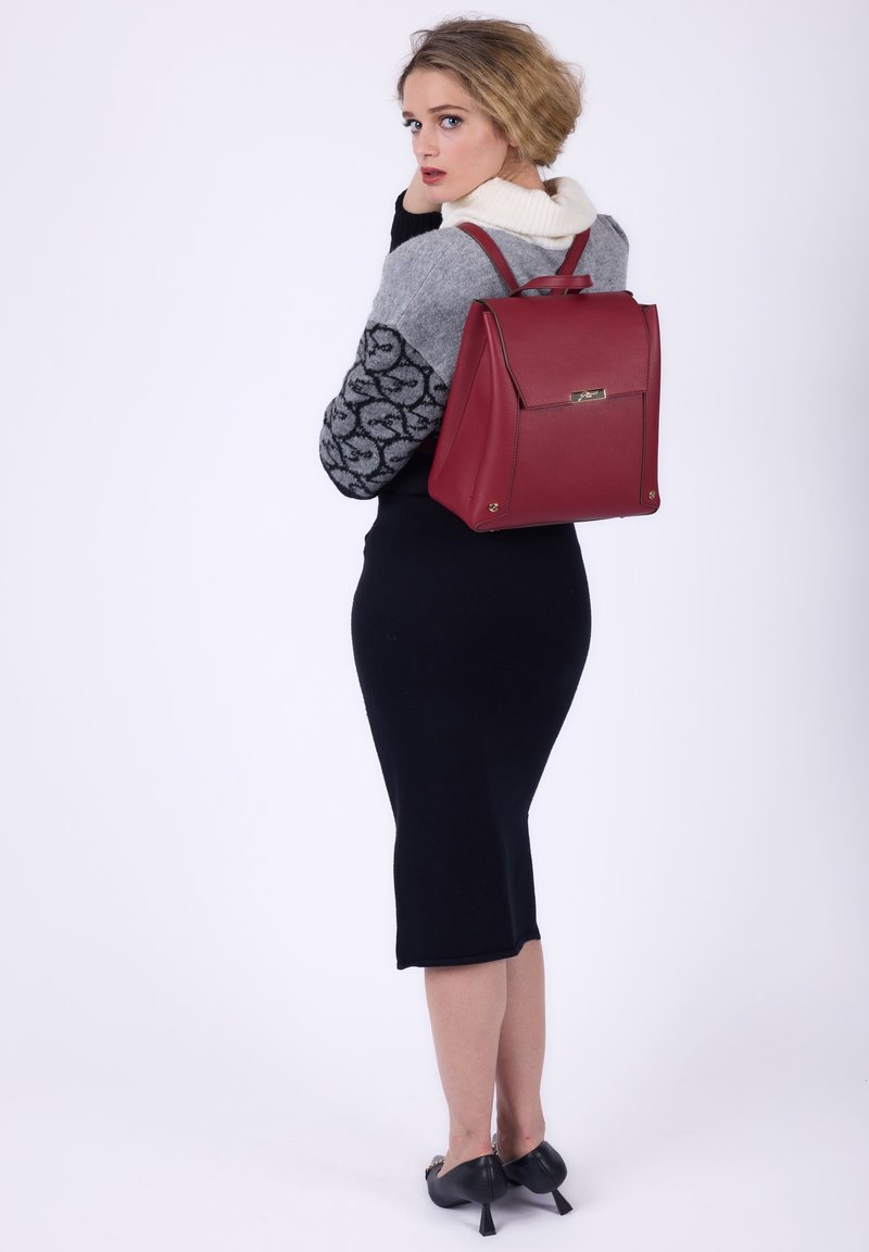 Donna in gonna nera e maglione grigio che indossa uno zaino rosso, volta le spalle con uno sfondo bianco semplice.