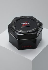 Black hexagonal metal tin with a silver band, featuring a top logo and embossed text, labeled "G-SHOCK," and noting water resistance.