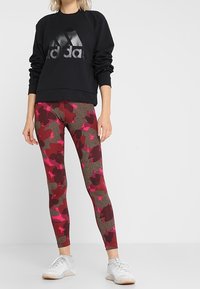 Sudadera negra con un logo de Adidas texturizado y brillante, y leggings estampados que presentan tonos de rojo, rosa y gris oscuro en un diseño de camuflaje.