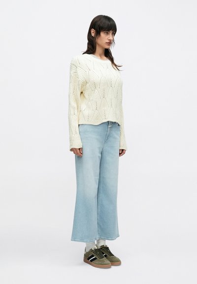 Γυναίκα που φοράει μπεζ πουλόβερ με πλέξη cable, ανοιχτόχρωμα τζιν με φαρδιές ελαστικές παντελόνες, λευκές κάλτσες και ελιά πράσινα αθλητικά παπούτσια, στέκεται μπροστά σε απλό λευκό φόντο.