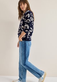 Dunkelblauer flauschiger Pullover mit hellgrauem Leopardenmuster, kombiniert mit hellblauen geraden Jeans. Das Model steht lässig mit den Händen in den Taschen.