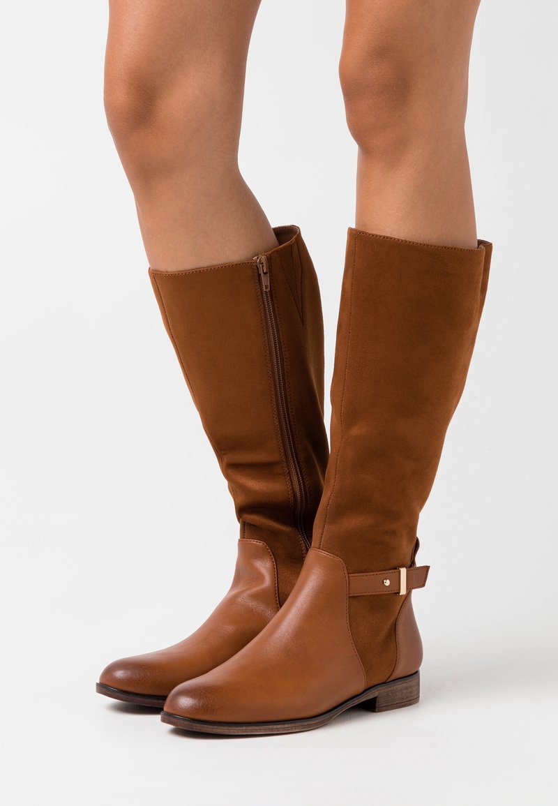 Bottes hautes jusqu'au genou en cuir marron à texture lisse, avec une fermeture éclair latérale et une sangle décorative ornée d'un accent doré.