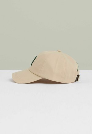 Cappellino da baseball beige con visiera rigida, caratterizzato da un logo verde sulla parte frontale e una patta regolabile sul retro. Tessuto strutturato.