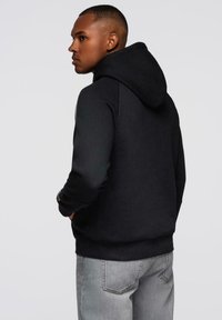 Mann mit kurzen Haaren, der einen schwarzen Kapuzenpullover und hellgraue Jeans trägt, nach links blickend vor einem schlichten hellen Hintergrund.