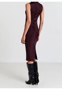 Femme portant une robe midi sans manches mauve foncé avec des côtés froncés et des bottes noires mi-hauteur jusqu'aux genoux, vue de dos sur un fond clair uni.