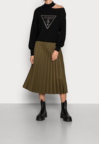Zwarte trui met een driehoekig logo, olijfgroene geplooide rok en zwarte enkellaarsjes met stevige zolen.