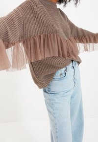 Brązowy dzianinowy sweter z warstwą przezroczystego tiulu na dole, noszony z jasnoniebieskimi spodniami wysokiej talii. Teksturowana tkanina i luźny krój.