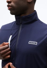 Άνδρας που φοράει σκούρο μπλε μπουφάν Calvin Klein, κουμπώνει την τσέπη με φερμουάρ στο στήθος με το ένα χέρι, ενώ στέκεται μπροστά σε λευκό φόντο.