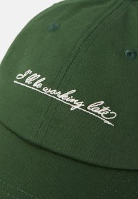 Grønn caps med hvit brodert tekst som sier "Jeg jobber sent." Nærbilde med fokus på teksturen i stoffet og broderidetaljer.