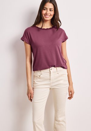 Kvinde smilende, iført plumfarvet kortærmet top og højtaljede off-white bukser, står mod en ensfarvet lys baggrund.