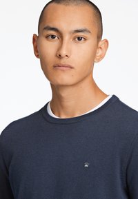 Pull marin avec un col rond et un col intérieur blanc. Logo en argent notable en forme de couronne sur la poitrine gauche. Matériau en coton avec une texture lisse.