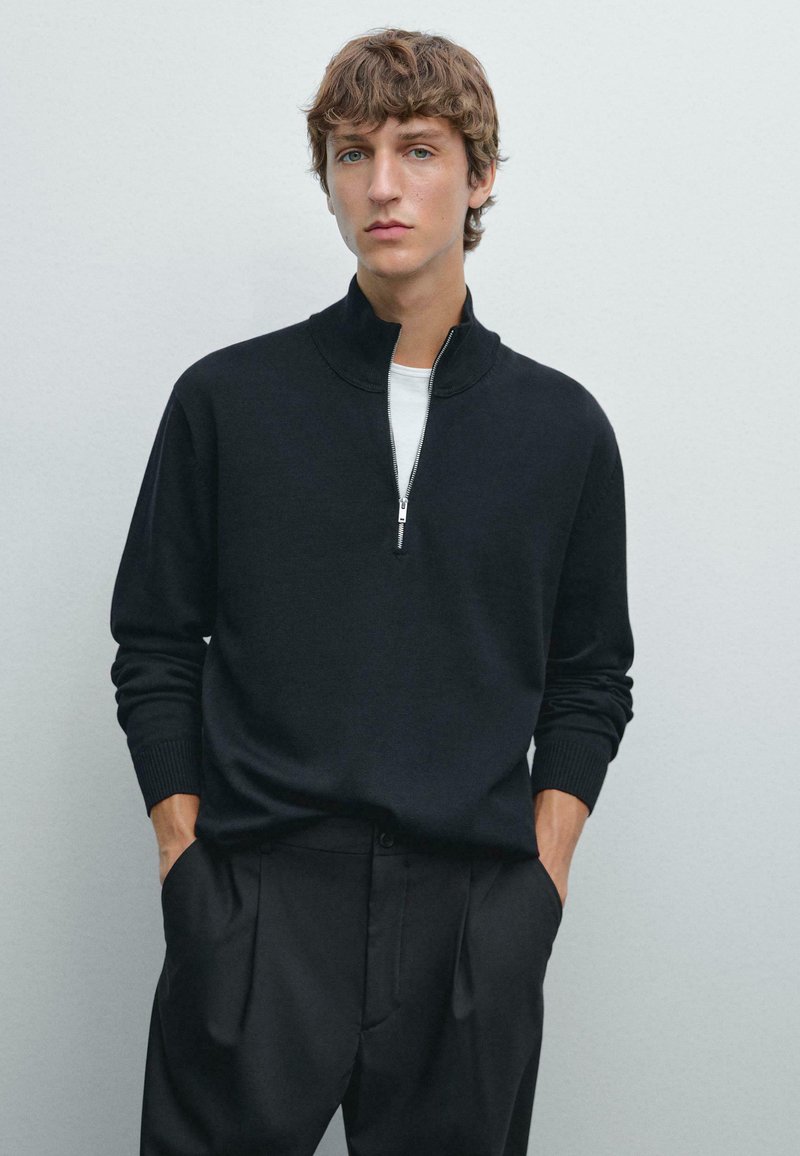 Maglione nero lavorato a maglia con colletto a mezza zip e polsini a coste, abbinato a pantaloni neri a vita alta. Texture liscia, vestibilità casual.