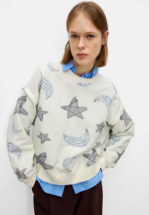 Felpa beige chiaro con motivi di stelle e lune grigi. Presenta un collo rotondo, maniche lunghe e una vestibilità comoda. L'orlo della camicia blu è visibile.