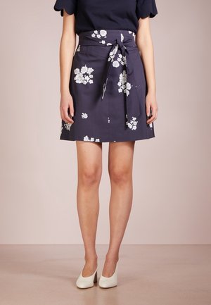 Femme portant une jupe bleu marine avec des motifs floraux blancs et une taille nouée, accompagnée de chaussures blanches à talons.