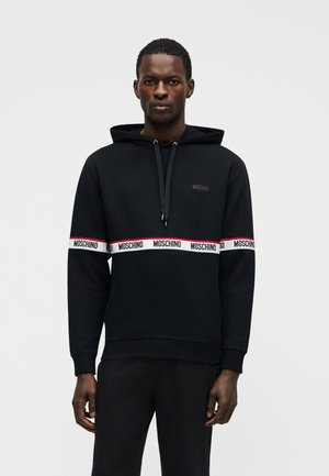 Homme portant un sweat à capuche noir Moschino avec un ruban logo blanc et rouge sur la poitrine et les manches, debout devant un fond clair uni.