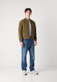 Olive green bomber jacket with a ribbed collar, paired with light blue denim jeans. White sneakers with grey accents complete the outfit.