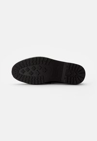 Semelle de chaussure en caoutchouc noir avec un motif texturé comportant des rainures et des sections surélevées pour une meilleure adhérence. Marque estampillée visible sur le côté.