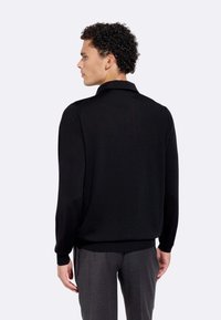 Pullover nero lavorato a maglia con colletto, maniche lunghe e polsini a coste. Materiale testurizzato con una finitura liscia. Indossato su pantaloni grigi.
