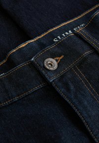 Tummansiniset slim-fit farkut, joissa on näkyviä oransseja ompeleita. Metallinen nappi, jossa on kaiverrus. Materiaali näyttää sileältä ja siinä on hienoinen kiilto.
