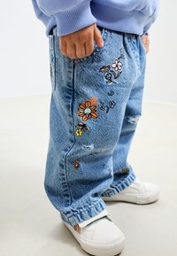 Világoskék farmernadrág virágos hímzéssel, kopott hatású részletekkel és gumírozott szegéllyel, fehér slip-on sportcipővel párosítva.