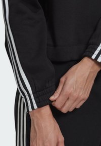 Svart sweatshirt med vita ränder längs ärmarna. Muddarna är samlade med elastik för en åtsittande passform. Materialet verkar mjukt och slätt.