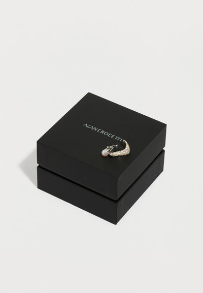 Μαύρο κουτί κοσμημάτων με ασημένιο λογότυπο, στη κορυφή του ένα χρυσόσκονιστο σκουλαρίκι σε σχήμα ημισελήνου διακοσμημένο με κρυστάλλους και με κρεμαστή λευκή πέρλα.