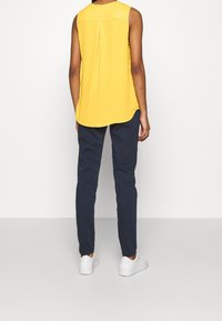 Persona che indossa una maglietta senza maniche gialla, pantaloni blu scuri e sneakers bianche, in piedi di fronte a un muro bianco semplice.