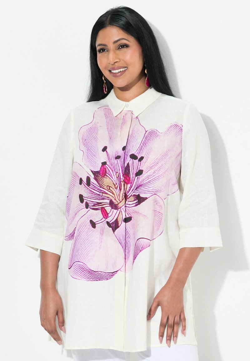Chemise en lin blanche ornée d'un grand motif florale rose, à boutons sur le devant et à manches trois-quarts. Accentuée de détails noirs dans la fleur.