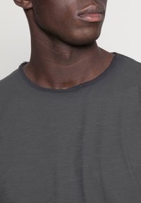 Primer plano de la parte inferior de la cara de una persona, cuello y parte superior del pecho, que lleva una camiseta de cuello redondo gris oscuro, mirando hacia un lado.
