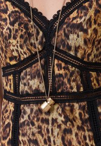 Vestido de estampado de leopardo com acabamento em renda preta, apresentando um decote em V e painéis estruturados. Um colar de pendente geométrico dourado pende abaixo.