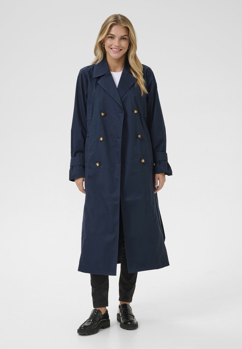 Marineblauwe dubbelgeknoopte trenchcoat met gouden knopen, lange mouwen en een rechte snit. Gedragen over een witte top en zwarte broek.