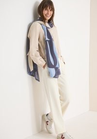 Femme appuyée contre un mur blanc, portant une veste beige clair, un pantalon crème, une écharpe bleue à motifs et des baskets blanches avec des accents rouges.