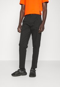 Pantaloni neri con vestibilità slim, caratterizzati da una vita piatta e passanti per cintura, abbinati a una maglietta arancione brillante e scarpe nere con cinturini.