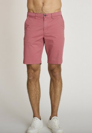 Homme portant un short rose jusqu'aux genoux et des baskets blanches, debout devant un fond clair uni.