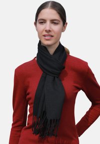 Écharpe noire en tissu doux, avec des extrémités frangées et un design épais enroulé. Portée sur un pull rouge.