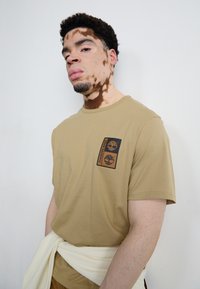Beige t-shirt met korte mouwen en een zwart Timberland logo-opdruk op de borst. Gladde stof, relaxte pasvorm, gecombineerd met een witte tailleband.