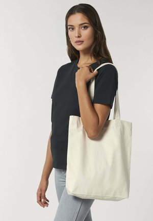 Jonge vrouw met lang haar, die een zwart t-shirt en lichte jeans draagt, houdt een grote simpele beige schoudertas over haar schouder tegen een effen achtergrond.