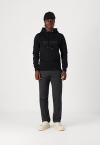 Sudadera negra con "ORIGINALS" impreso en letras oscuras, pantalones grises, zapatillas beige y una gorra negra, de pie sobre un fondo neutral.