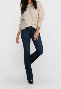 Jean en denim foncé à coupe droite, associé à un pull texturé beige clair. La tenue se caractérise par un minimum d'accessoires et une coupe décontractée.