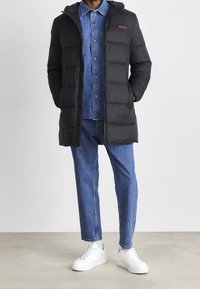 Schwarzer Daunenmantel mit Kapuze, horizontaler Steppung und gesticktem Logo. Getragen über einem Jeanshemd, kombiniert mit blauen Jeans und weißen Turnschuhen.